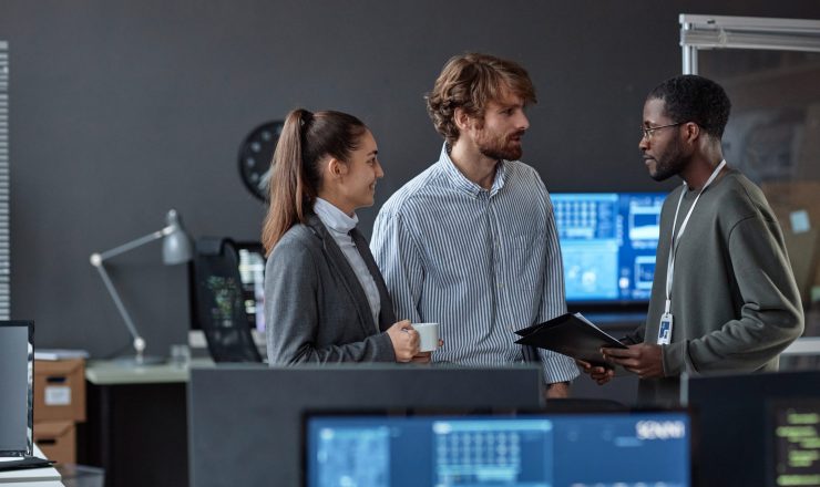 Over desktop view of three young people chatting standing in office with multiple computers copy space